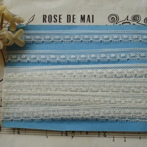 May include: A collection of off-white lace trims on a light blue card. The lace features various delicate patterns, including floral and geometric designs. The text "ROSE DE MAI" is visible at the top. The image is set against a backdrop of sheet music and flowers.