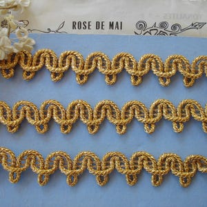 May include: Close-up of three rows of gold-colored decorative trim with a looped design. The trim is made of a twisted, rope-like material. The background is a light blue surface with the text "ROSE DE MAI".