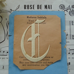 May include: A vintage paper craft kit for making custom initials. The kit includes a white paper letter 'E' with a decorative script font. The text on the paper reads "Reform Initials, Wash It, Easy to Embroider, Can be combined in letters or monograms, Buy your own initials and make any combination you like."