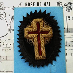 May include: A decorative cross emblem featuring a deep red velvet center bordered by intricate gold metallic thread embroidery. The cross is mounted on a black pleated fabric oval base with a pointed, starburst-like edge, creating a textured effect.
