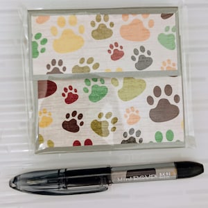 Puede incluir: Dos tarjetas con un diseño de huellas de patas multicolores, empaquetadas en plástico transparente. Debajo hay un bolígrafo negro. El bolígrafo tiene el texto "Mini RSVP BK91 MED".