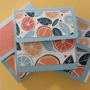 May include: A collection of small, square paper boxes with a citrus fruit design. The boxes display orange, red, and blue citrus slices and leaves on a white backdrop, with light blue borders. The background is yellow.