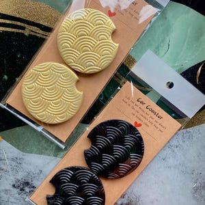May include: Two sets of car coasters in clear plastic packaging. One set is gold with a wave pattern, the other is black with a similar design. The packaging includes the text "Car Coaster".