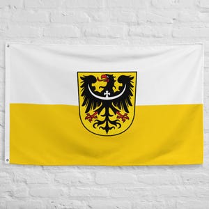 May include: A rectangular flag featuring two horizontal stripes of white on the top and yellow on the bottom. Centered on the flag is a shield displaying a black eagle with a red beak and claws, featuring a white crescent moon and cross.