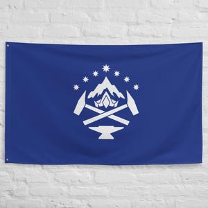 May include: A blue flag with a white emblem featuring a mountain range, stars, hammers, and an anvil. The flag is rectangular and hangs against a white brick wall.