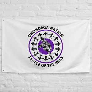 Könnte beinhalten: Weiße Flagge mit dem schwarzen Schriftzug "ONONDAGA NATION", der einen lila Kreis mit einem indianischen Profil und den Worten "PEOPLE OF THE HILLS" umgibt. Die Flagge hat Ösen zum Aufhängen.