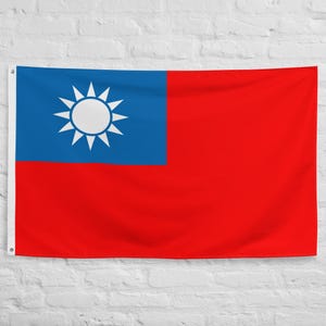 May include: A rectangular flag featuring the flag of Taiwan. The flag has a red field with a blue canton in the upper left corner. The canton contains a white sun with twelve rays. The flag is hanging against a white brick wall.