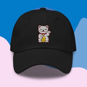 Può includere: Cappellino da baseball nero con un Maneki-Neko bianco ricamato, un gatto portafortuna giapponese, che tiene una moneta d'oro. Il gatto ha le orecchie rosa e un collare rosa con un campanello.