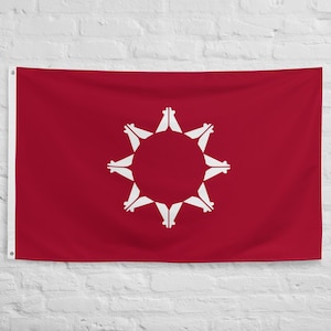 May include: A red flag with a white emblem in the center. The emblem is a circle with white shapes radiating outwards. The flag is hanging against a white brick wall.