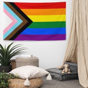 Può includere: Una bandiera rettangolare con il design della bandiera Progress Pride. Presenta uno sfondo arcobaleno con strisce nere, marroni, azzurre, rosa e bianche, e un chevron nero. La bandiera è appesa a una parete bianca.