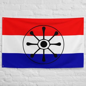 May include: A rectangular flag with horizontal stripes of red, white, and blue. A black emblem resembling a wheel with eight spokes is centered on the flag. The flag is hanging against a white brick wall.