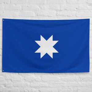 Puede incluir: Una bandera azul con una estrella blanca de ocho puntas en el centro. La bandera es rectangular y cuelga contra una pared de ladrillos blancos. La estrella es de color blanco brillante y destaca sobre el fondo azul.