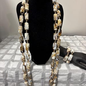 Könnte beinhalten: Ein zweiteiliges Halsketten- und Armband-Set. Der Schmuck besteht aus großen, rechteckigen Perlen in Weiß-, Braun- und Beigetönen. Die Halskette wird auf einer schwarzen Schaufensterpuppe und das Armband auf einer schwarzen Hand präsentiert.