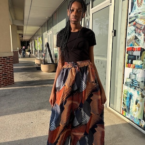 Puede incluir: Una persona lleva una camiseta negra y pantalones de pernera ancha con un estampado abstracto colorido en tonos naranja, azul y blanco. Los pantalones tienen cintura con cordón. La persona lleva zapatillas deportivas blancas y negras.