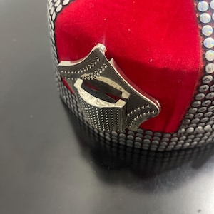 Peut inclure: Un chapeau en velours rouge avec des ornements argentés. Le chapeau comporte une pièce décorative en métal argenté sur le côté. Le chapeau est orné de strass argentés et de petits clous argentés.