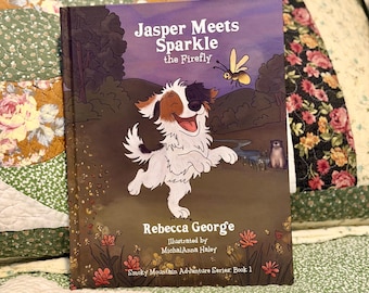May include: A hardcover children's book titled Jasper Meets Sparkle the Firefly by Rebecca George, illustrated by MichalAnna Haley. The cover features a white and brown puppy jumping in a field with a firefly, a small stream, and pink flowers.