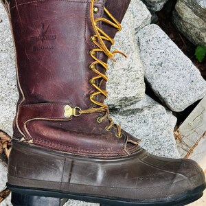 May include: A dark brown leather boot with a rubber lower shell and a black lugged sole. The upper features yellow laces threaded through brass eyelets and hooks. The side is embossed with the text LaCrosse and Big Mountain branding.