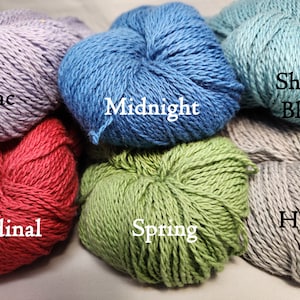 May include: Close-up of several skeins of yarn in various colors, including lilac, midnight blue, cardinal red, spring green, and haze gray. Each skein is labeled with its color name in white text. The yarn appears soft and textured.