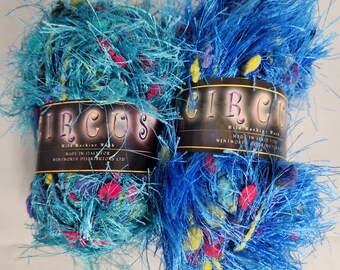 May include: Two skeins of vibrant blue yarn with a fluffy, textured appearance. The yarn is speckled with colorful accents of pink, yellow, and purple. Each skein has a label that reads "CIRCUS".