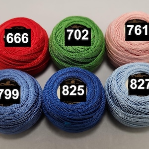 以下が含まれることがあります： さまざまな色の刺繍糸の糸巻きが6つあります。糸巻きには、666、702、761、799、825、827の番号が付けられています。