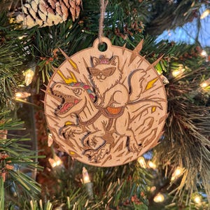 May include: Wooden Christmas ornament featuring a cat wearing sunglasses and a crown riding a dinosaur. The ornament is round with a light brown finish and a hole for hanging. Lightning bolt designs are etched around the cat and dinosaur.