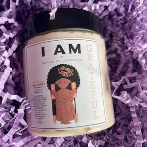 May include: A white jar with a black lid and a label that says "I AM by HOUSE OF LAVANDER" and "GRATEFUL". The jar contains a body butter called "Cies Mango" with a mango scent. The label also includes a cartoon illustration of a woman with a brush and the words "LAVANDER LIPSTICK". The jar is 8 oz.