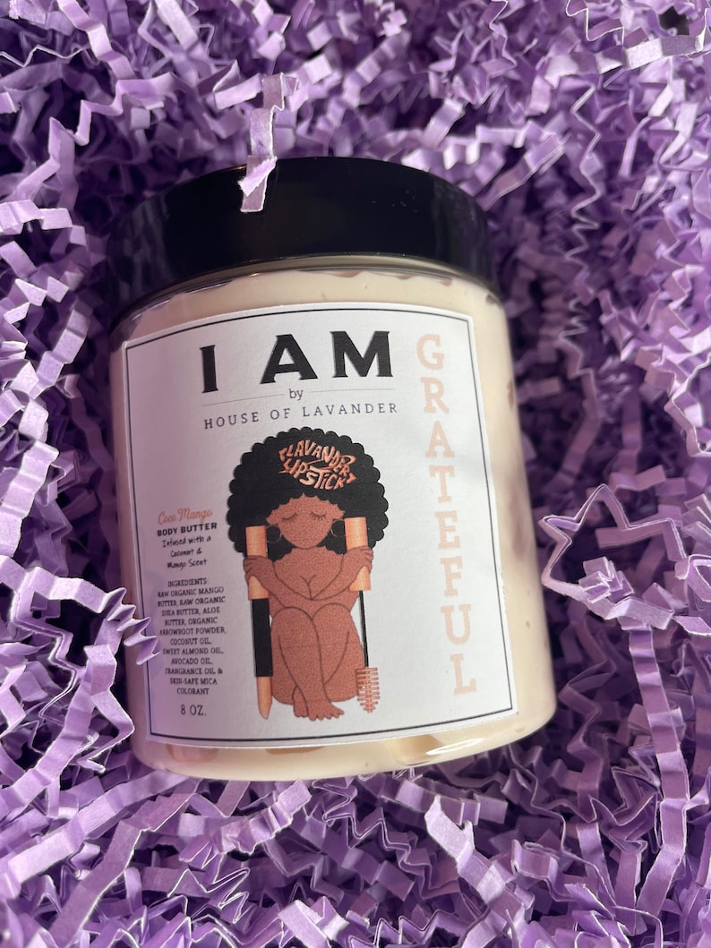 May include: A white jar with a black lid and a label that says "I AM by HOUSE OF LAVANDER" and "GRATEFUL". The jar contains a body butter called "Cies Mango" with a mango scent. The label also includes a cartoon illustration of a woman with a brush and the words "LAVANDER LIPSTICK". The jar is 8 oz.