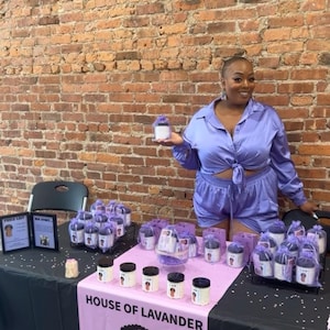 May include: A woman wearing a purple satin outfit stands behind a table displaying jars of lavender-scented products. The table is covered with a black tablecloth and a pink tablecloth with the words "House of Lavander" and a cartoon image of a woman. The jars are arranged in rows on the table and are labelled with the words "I Am" and "Lavender Supreme".