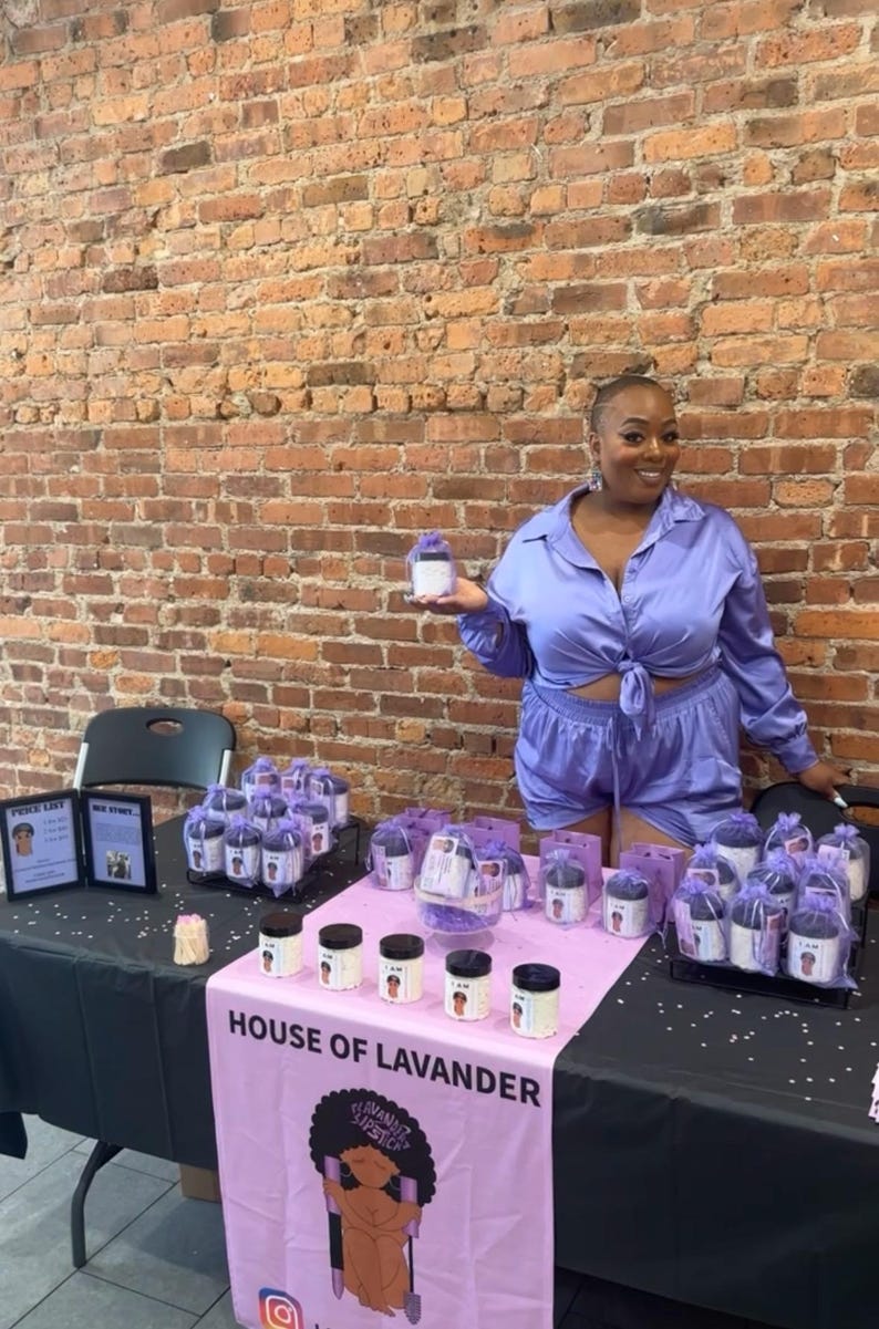 May include: A woman wearing a purple satin outfit stands behind a table displaying jars of lavender-scented products. The table is covered with a black tablecloth and a pink tablecloth with the words "House of Lavander" and a cartoon image of a woman. The jars are arranged in rows on the table and are labelled with the words "I Am" and "Lavender Supreme".