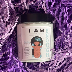 May include: A white jar of body butter with a black lid and a colourful illustration of a woman holding a lipstick. The jar is labelled "I AM ENOUGH" by "HOUSE OF LAVENDER". The body butter is infused with mango, shea butter, aloe, coconut oil, sweet almond oil, avocado oil, and fragrance oil. The jar is 8 oz.