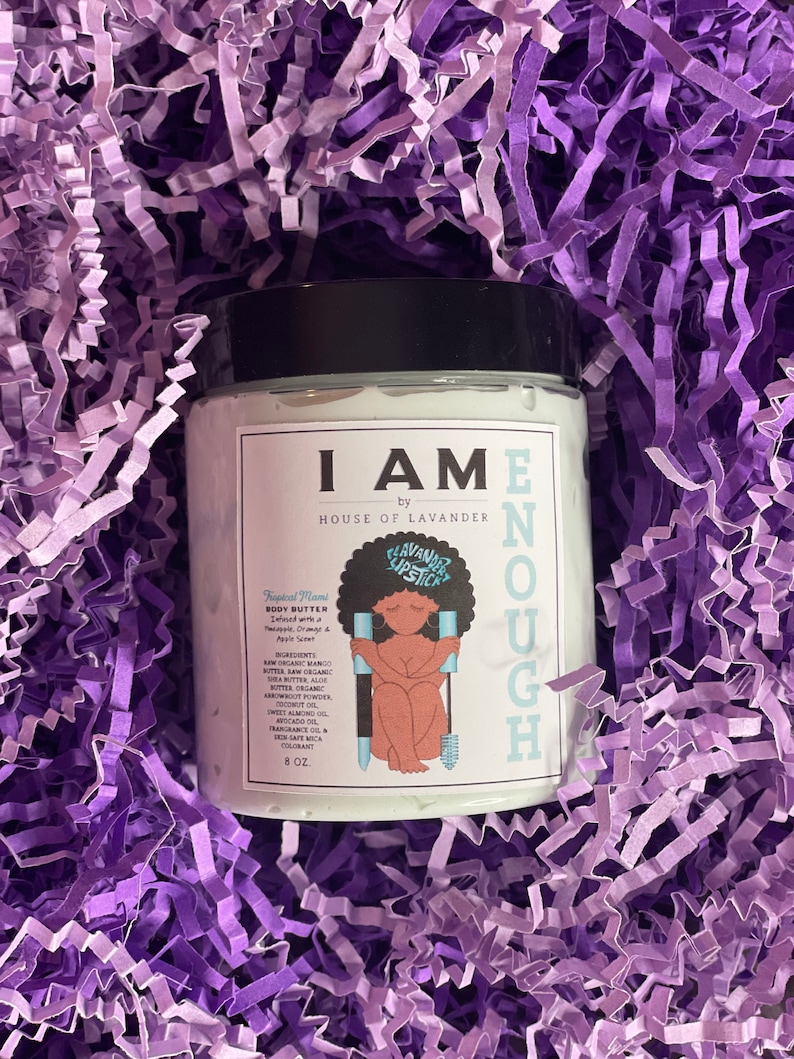 May include: A white jar of body butter with a black lid and a colourful illustration of a woman holding a lipstick. The jar is labelled "I AM ENOUGH" by "HOUSE OF LAVENDER". The body butter is infused with mango, shea butter, aloe, coconut oil, sweet almond oil, avocado oil, and fragrance oil. The jar is 8 oz.