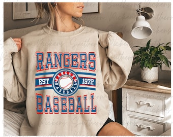 Puede incluir: Una sudadera beige con un diseño gráfico rojo y azul que dice "RANGERS BASEBALL EST 1972" con una pelota de béisbol y estrellas en un círculo.