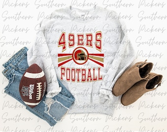 Pode incluir: Uma camisa de moletom cinza com as palavras "49ERS FOOTBALL" em letras douradas e vermelhas. A camisa de moletom está colocada sobre uma superfície branca com uma bola de futebol, uma calça jeans azul e botas marrons.