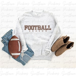 Puede incluir: Una sudadera gris con el texto "FOOTBALL SEASON" en letras marrones. La sudadera est&aacute; colocada sobre una superficie blanca con una pelota de f&uacute;tbol marr&oacute;n, unos vaqueros azules y unas botas marrones de tobillo.