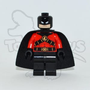 Peut inclure: Une figurine Lego d'un personnage de super-héros. La figurine a une tête noire et des jambes noires. Le torse est rouge avec un motif or et noir. Une cape noire est drapée sur les épaules. La figurine est debout.