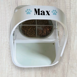 Op de afbeelding: Witte automatische huisdiervoederdispenser met twee compartimenten voor droogvoer. De dispenser heeft een doorzichtig plastic deksel en een witte basis met de naam "Max" en twee blauwe voetafdrukken.