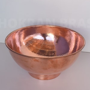 May include: A hammered copper bowl with a wide, rounded shape and a small, circular base. The bowl has a shiny, reflective surface and a warm, reddish-brown color. The bowl is empty and sits on a white surface.