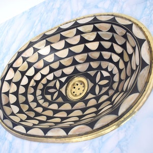 Op de afbeelding: Een ovale wastafel met een zwart-wit geometrisch patroon. De wastafel is gemaakt van een materiaal dat lijkt op bot of schelp. De wastafel heeft een messing rand en een afvoer in het midden.