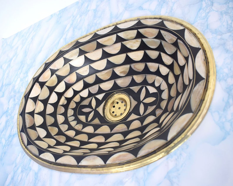 Op de afbeelding: Een ovale wastafel met een zwart-wit geometrisch patroon. De wastafel is gemaakt van een materiaal dat lijkt op bot of schelp. De wastafel heeft een messing rand en een afvoer in het midden.