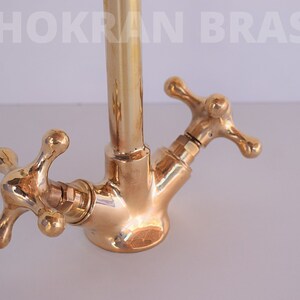 May include: Close-up of a gold-toned brass kitchen faucet with two cross-shaped handles. The faucet has a tall, cylindrical spout and is designed in a traditional style.