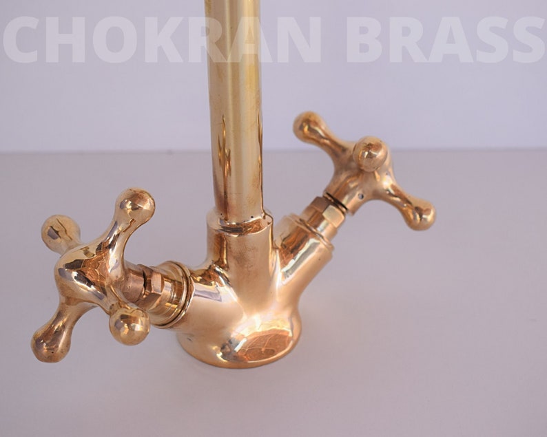 May include: Close-up of a gold-toned brass kitchen faucet with two cross-shaped handles. The faucet has a tall, cylindrical spout and is designed in a traditional style.