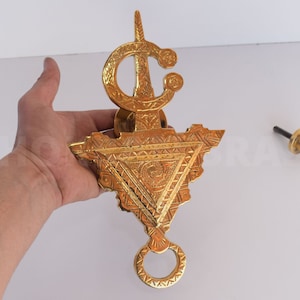 Puede incluir: Un llamador de puerta de metal dorado con intrincados diseños geométricos. El llamador tiene forma triangular con un anillo en la parte inferior y una parte superior decorativa con un remate puntiagudo.