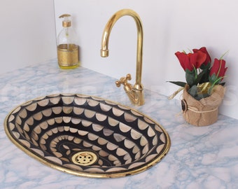 May include: A black and white oval sink with a gold rim and a brass faucet. The sink is decorated with a pattern of overlapping semicircles. The sink is sitting on a white countertop.