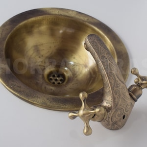 Peut inclure: Un évier en laiton avec un robinet orné. L'évier est rond et présente un motif décoratif autour du bord. Le robinet a un bec incurvé et deux poignées en forme de croix.