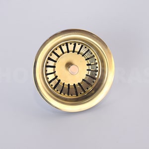 May include: A gold-colored metal sink strainer with a circular design and a central hole. The strainer has a series of small holes around the edge.