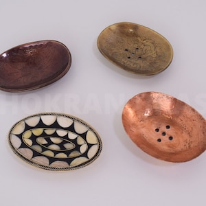 Peut inclure: Quatre porte-savons ovales en cuivre, laiton et matériaux décoratifs. L'un est de couleur cuivre avec six trous de drainage. Un autre est en laiton avec un motif floral gravé. Le troisième a un design incrusté noir et blanc. Le dernier est brun foncé avec un motif gravé.