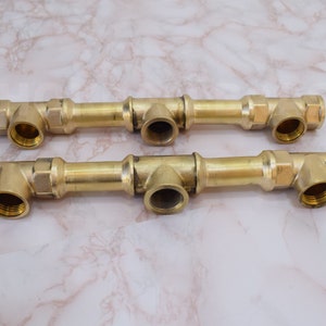 Może przedstawiać: Dwa rury wodociągowe z mosiądzu z wieloma złączami. Każda rura ma trzy złącza, jedno na każdym końcu i jedno na środku. Złącza są wszystkie w kształcie litery T.