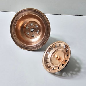 May include: Two copper sink strainers, one with a larger diameter and a decorative pattern, and the other with a smaller diameter and a more simple design.