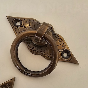 Peut inclure: Une poignée de porte en laiton avec un anneau décoratif et une plaque en forme de diamant ornée de motifs floraux complexes.