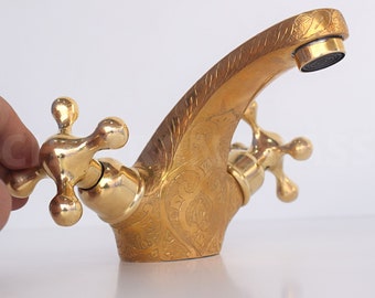 May include: A gold-toned faucet with intricate engraved designs. The faucet has two cross-shaped handles.